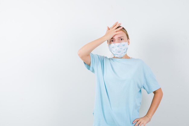 Junge Dame im T-Shirt, Maske, die Hand auf die Stirn hält und vergesslich aussieht