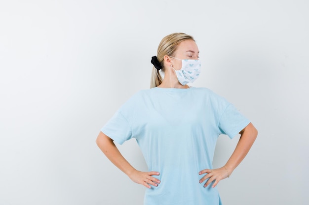 Junge Dame im T-Shirt, Maske, die mit den Händen auf der Taille posiert und selbstbewusst aussieht