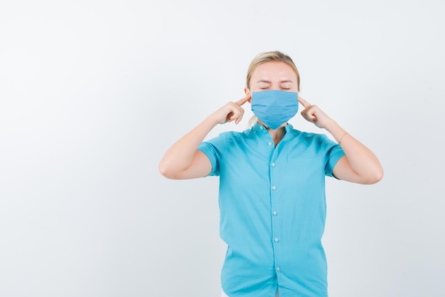 Junge Dame im T-Shirt, Maske, die Ohren mit den Fingern verstopft und friedlich aussieht