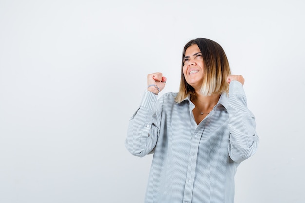 Junge Dame im übergroßen Hemd, die Gewinnergeste zeigt und glücklich aussieht, Vorderansicht.