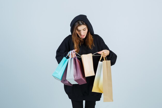 Junge Dame im übergroßen Kapuzenpulli, Hosen, die in Pakete schauen und erstaunt schauen, Vorderansicht.