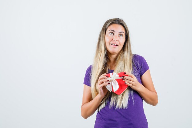 Junge Dame im violetten T-Shirt hält Geschenkbox und schaut fröhlich, Vorderansicht.
