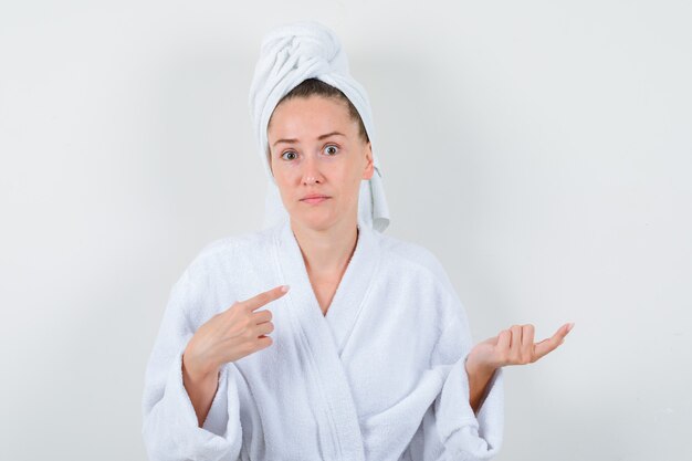 Junge Dame im weißen Bademantel, Handtuch zeigt zur Seite und sieht unentschlossen aus, Vorderansicht.