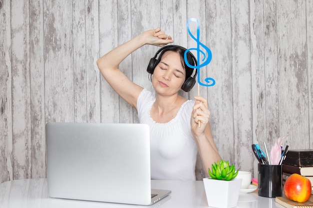 junge Dame im weißen Hemd, das blaue Note hält, die Musik durch schwarze Kopfhörer auf Tisch zusammen mit grünen Pflanzenstiften auf Grau hört