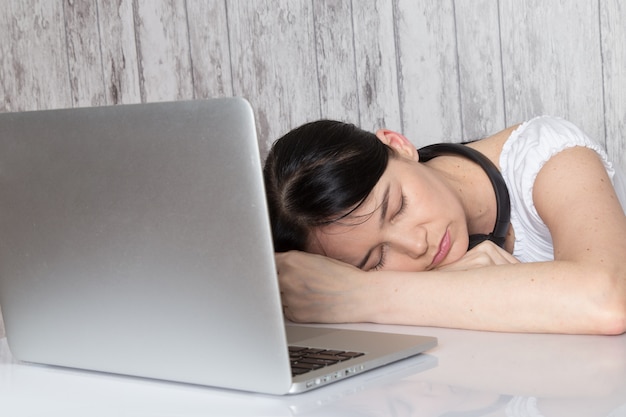 junge Dame im weißen Hemd mit schwarzen Ohrenschützern schlief vor Laptop auf Grau ein