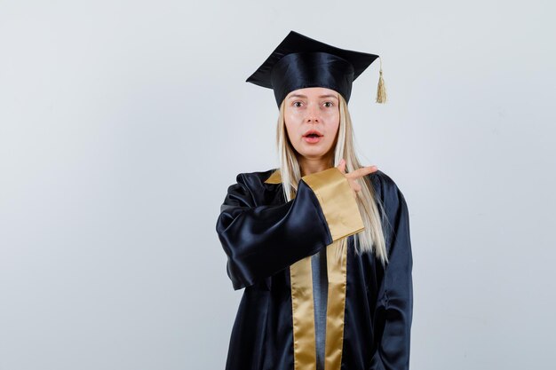 Junge Dame in akademischer Kleidung, die zur Seite zeigt und erstaunt aussieht