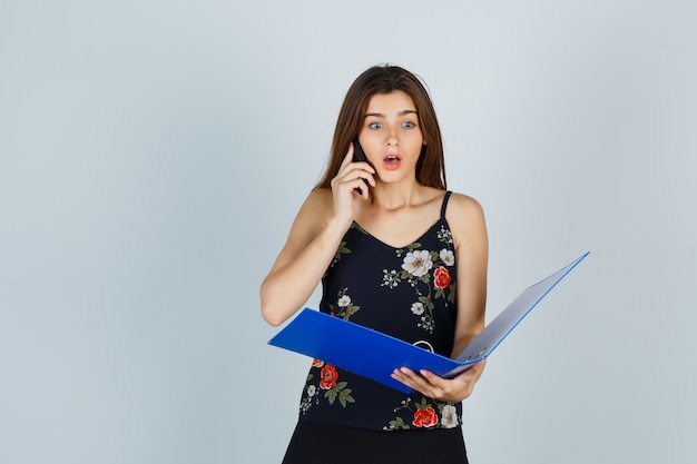 Junge Dame in Bluse, die Ordner hält, mit dem Handy spricht und schockiert aussieht, Vorderansicht.