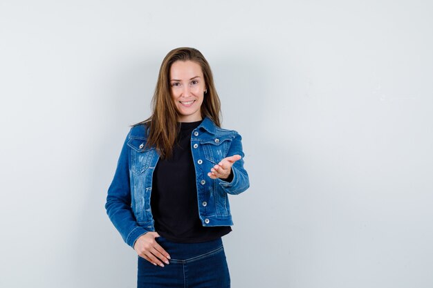 Junge Dame in Bluse, Jacke, Jeans, die die Hand ausstreckt, um etwas zu zeigen und selbstbewusst auszusehen