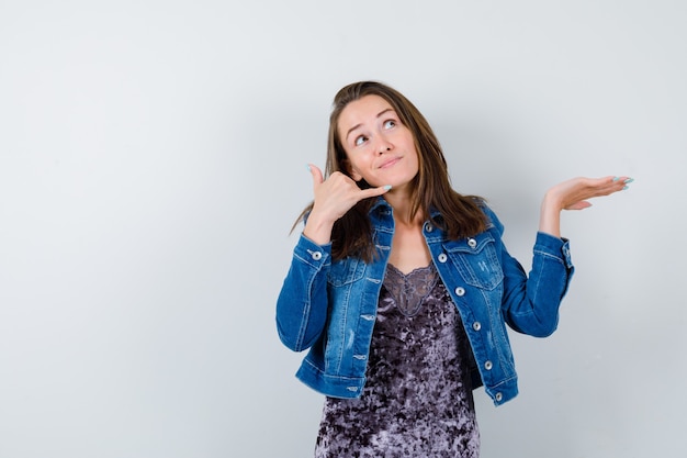 Junge Dame in Bluse, Jeansjacke, die Telefongeste zeigt und süß aussieht, Vorderansicht.