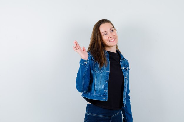 Junge Dame in Bluse winkt mit der Hand zur Begrüßung und sieht fröhlich aus, Vorderansicht.