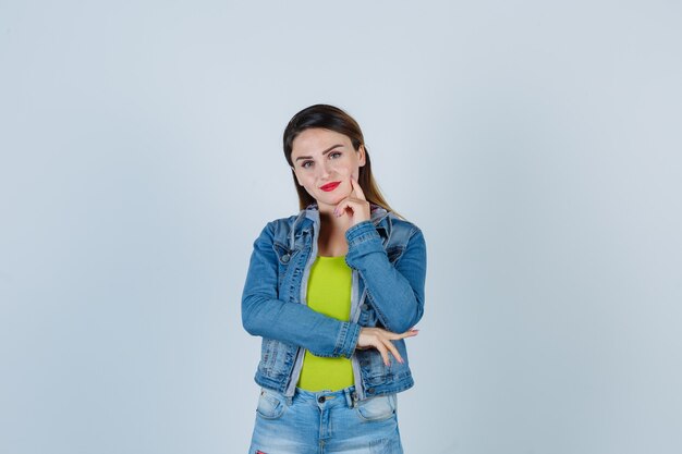 Junge Dame in Denim-Outfit, die das Kinn auf der Hand stützt, den Finger auf die Wange hält und positiv aussieht, Vorderansicht.