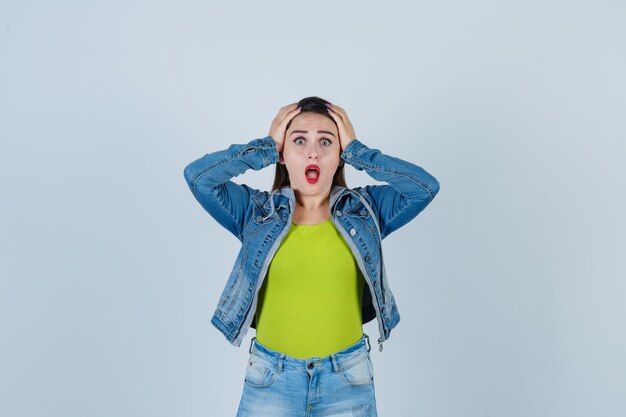 Junge Dame in Denim-Outfit, die die Hände auf den Kopf hält und aufgeregt aussieht, Vorderansicht.