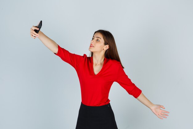 Junge Dame in der roten Bluse, Rock, der selfie nimmt, während Arm streckend und hübsch aussieht