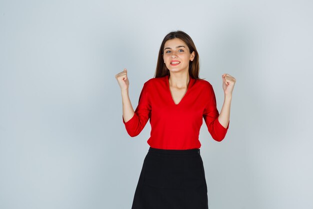 Junge Dame in der roten Bluse, Rock, der Siegergeste zeigt und glückselig aussieht