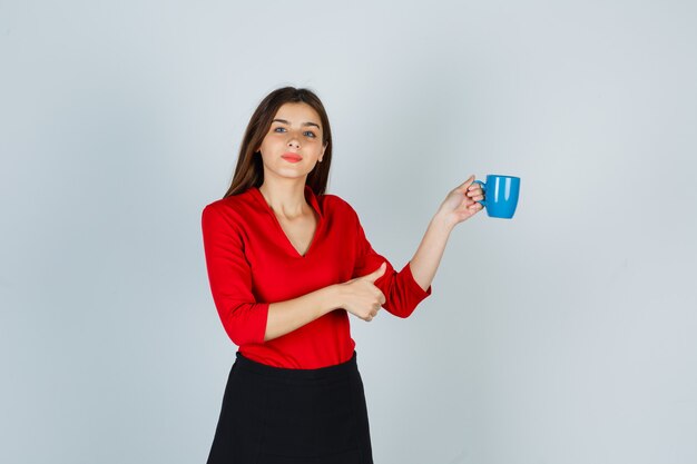 Junge Dame in der roten Bluse, Rock, der Tasse hält, während Daumen oben zeigt und zuversichtlich schaut