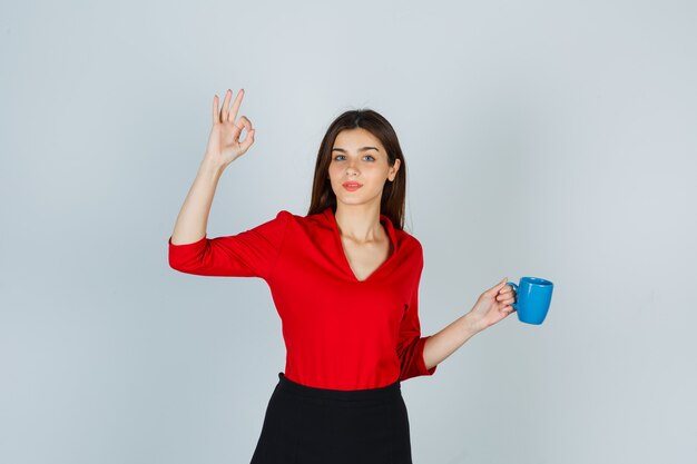 Junge Dame in der roten Bluse, Rock, der Tasse hält, während ok Geste zeigt