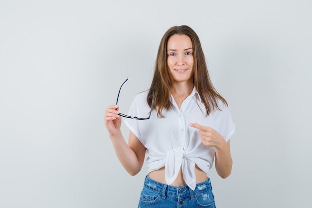 Junge Dame in der weißen Bluse, die auf Brille zeigt, Vorderansicht.