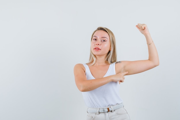 Kostenloses Foto junge dame in der weißen bluse, die auf ihre armmuskeln zeigt und konzentriert schaut