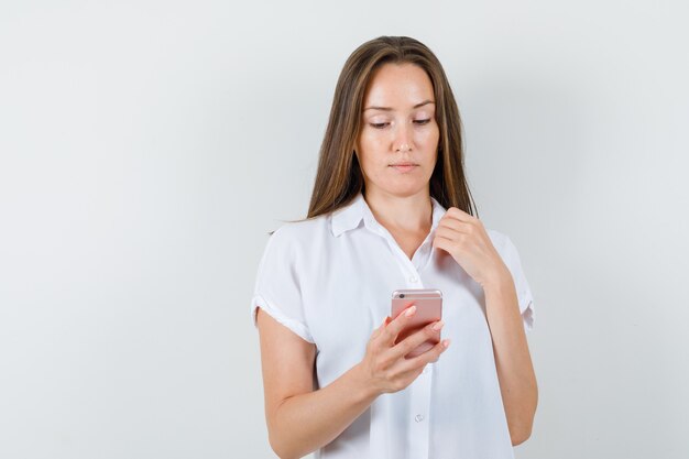 Junge Dame in der weißen Bluse, die Telefon betrachtet und konzentriert schaut