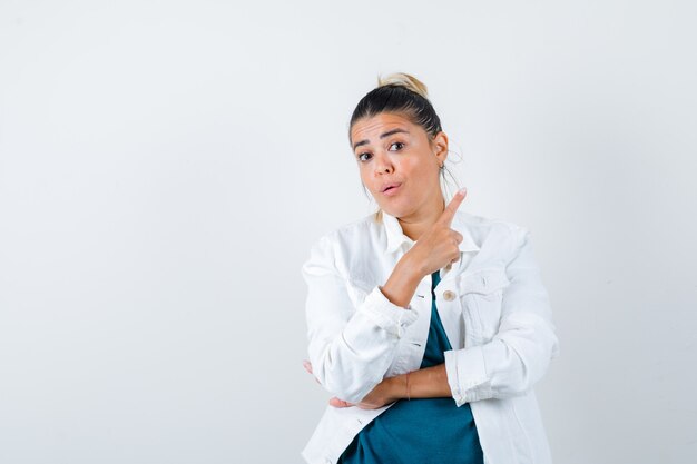 Junge Dame in der weißen Jacke zeigt auf die obere rechte Ecke und sieht verwundert aus, Vorderansicht.