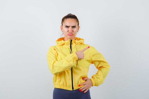 Junge Dame in gelber Jacke, die auf die obere rechte Ecke zeigt und ernst aussieht, Vorderansicht.