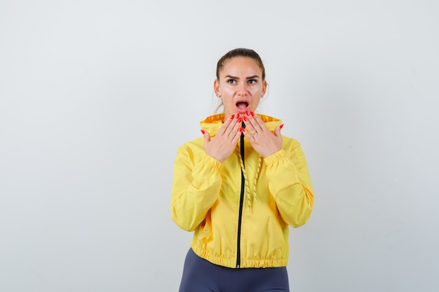 Junge Dame in gelber Jacke mit Händen in der Nähe des Mundes und verwirrt, Vorderansicht.