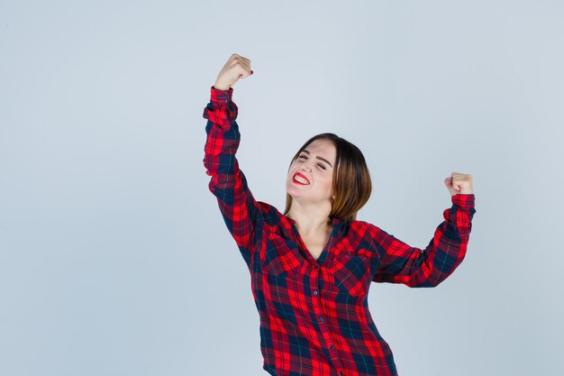 Junge Dame in kariertem Hemd, die Gewinnergeste zeigt und glückselig aussieht, Vorderansicht.