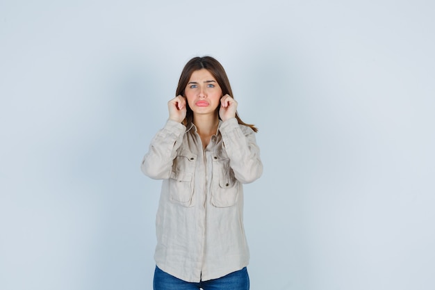 Junge Dame in lässigen Jeans, die Ohrläppchen ziehen, die Lippen schmollen und unzufrieden aussehen, Vorderansicht.