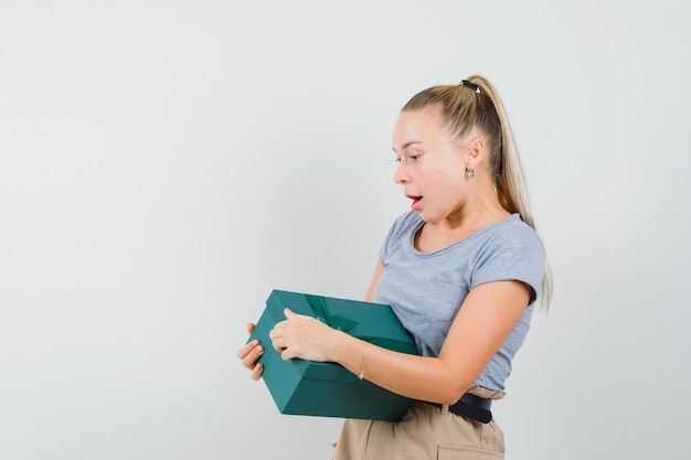 Junge Dame in T-Shirt und Hose, die versucht, Geschenkbox zu öffnen und neugierig schaut,