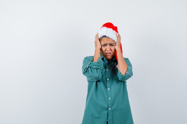 Junge Dame in Weihnachtsmütze, Hemd, das den Kopf mit den Händen umklammert und traurig aussieht, Vorderansicht.