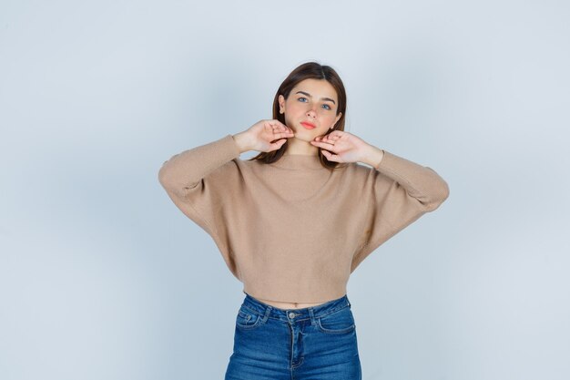 Junge Dame mit den Händen unter dem Kinn in beigefarbenem Pullover, Jeans und hübschem Aussehen. Vorderansicht.