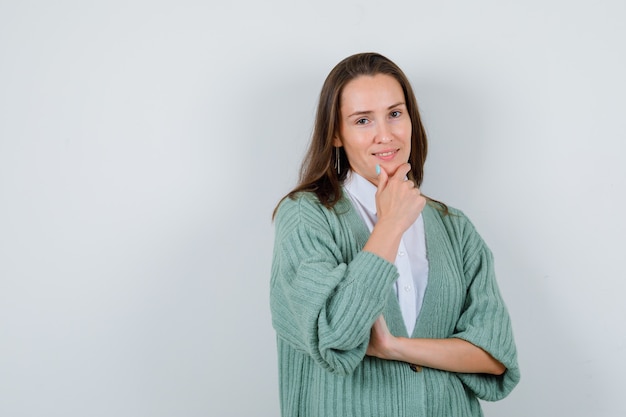 Junge Dame mit Hand am Kinn in Hemd, Strickjacke und zufrieden. Vorderansicht.