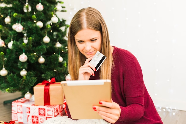 Junge Dame mit Tablette und Plastikkarte nahe Geschenkboxen und Weihnachtsbaum