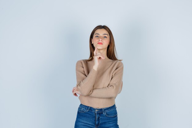Junge Dame posiert, während sie den Finger am Kinn in beigefarbenem Pullover, Jeans hält und selbstbewusst aussieht, Vorderansicht.