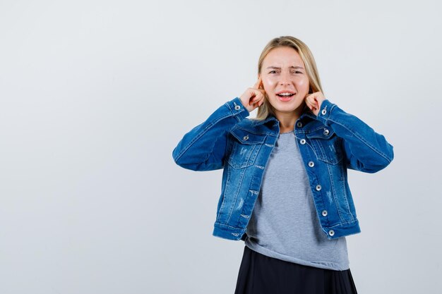 Junge Dame steckt Ohren mit Fingern in T-Shirt, Jeansjacke, Rock und sieht verwirrt aus