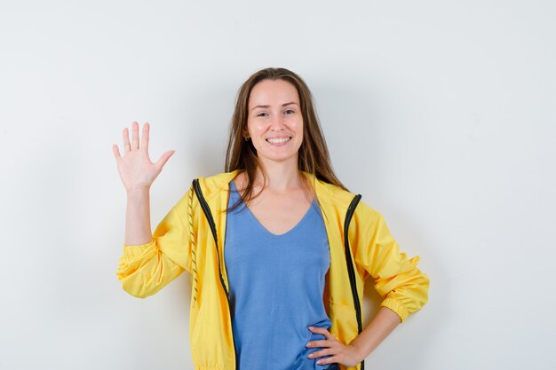 Junge Dame winkt mit der Hand zum Gruß in T-Shirt, Jacke und sieht entzückend aus. Vorderansicht.