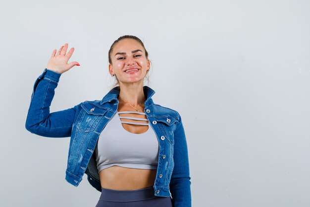 Junge Dame winkt mit der Hand zum Gruß in Top, Jeansjacke und sieht fröhlich aus. Vorderansicht.