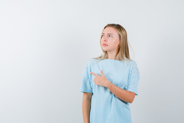 Junge Dame zeigt auf die linke Seite im T-Shirt und sieht zögernd aus. Vorderansicht.