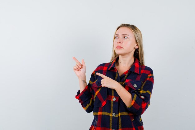 Junge Dame zeigt auf die obere linke Ecke im karierten Hemd und schaut zögernd, Vorderansicht.