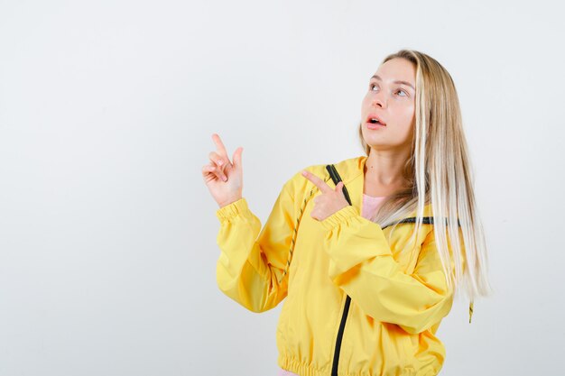 Junge Dame zeigt auf die obere linke Ecke in T-Shirt, Jacke und sieht verwirrt aus