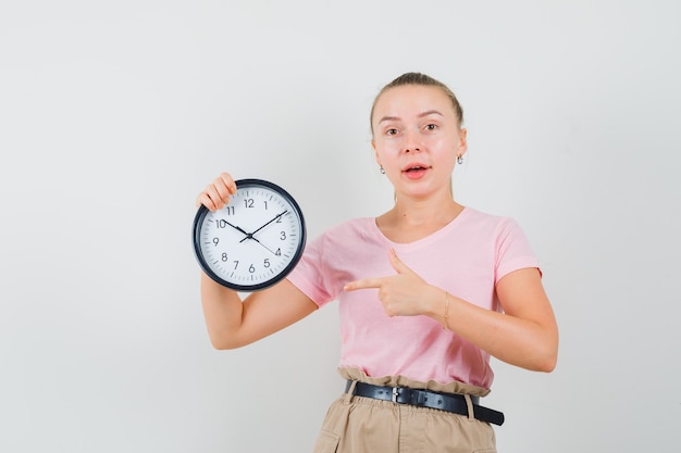 Junge Dame zeigt auf Wanduhr in T-Shirt und Hose und sieht positiv aus