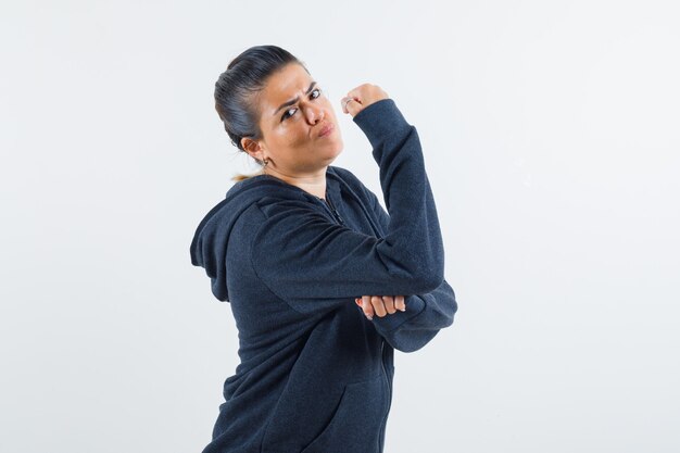 Junge Dame zeigt ihre Armmuskeln in Jacke und sieht streng aus. Vorderansicht.