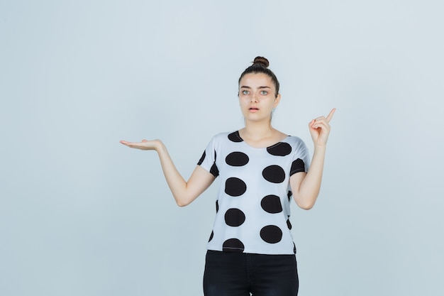 Junge Dame zeigt nach oben, während sie etwas in T-Shirt, Jeans zeigt und erstaunt, Vorderansicht schaut.