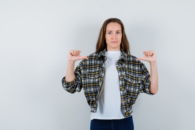 Junge Dame zeigt sich mit Daumen in T-Shirt, Jacke, Jeans und sieht stolz aus. Vorderansicht.