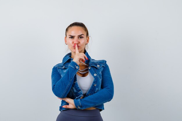 Junge Dame zeigt Stille Geste in Top, Jeansjacke und sieht ernst aus. Vorderansicht.