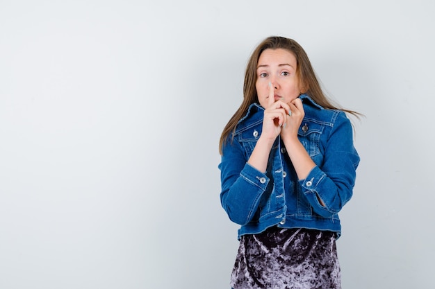 Junge Dame zeigt Stille Geste, zieht Kragen im Gesicht in Kleid, Jeansjacke und sieht verängstigt aus. Vorderansicht.