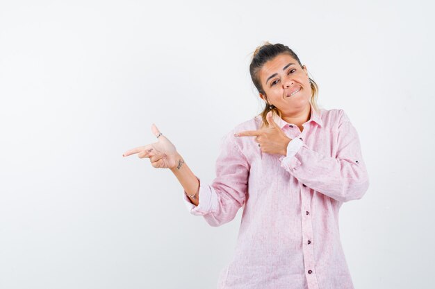 Junge Dame zeigt zur Seite, beißt sich auf die Lippe im rosa Hemd und sieht fröhlich aus