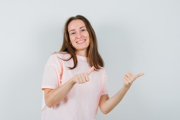 Junge Dame zeigt zur Seite mit Daumen hoch in rosa T-Shirt und sieht munter aus