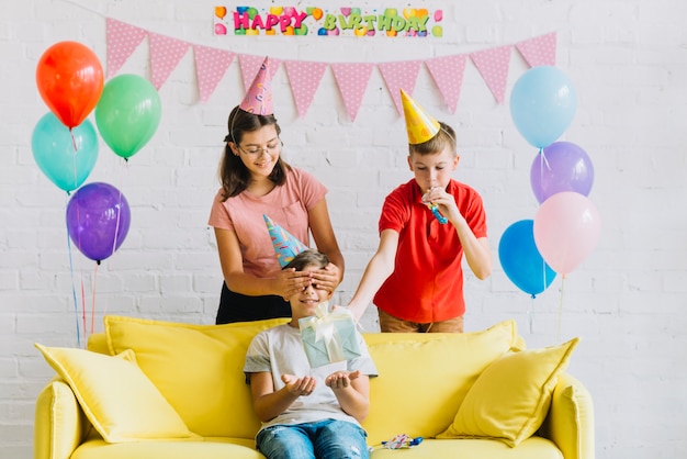 Junge, der auf dem Sofa sitzt, das Überraschungsgeburtstagsgeschenk von seinen Freunden empfängt