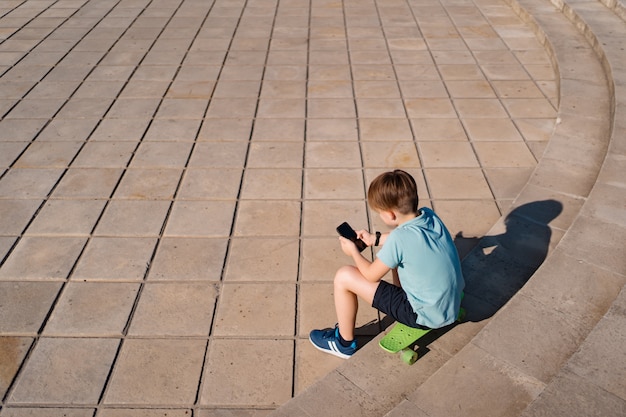 Junge, der auf Treppe mit Smartphone in seiner Hand und grünem Penny Board sitzt und lustige Videos sieht